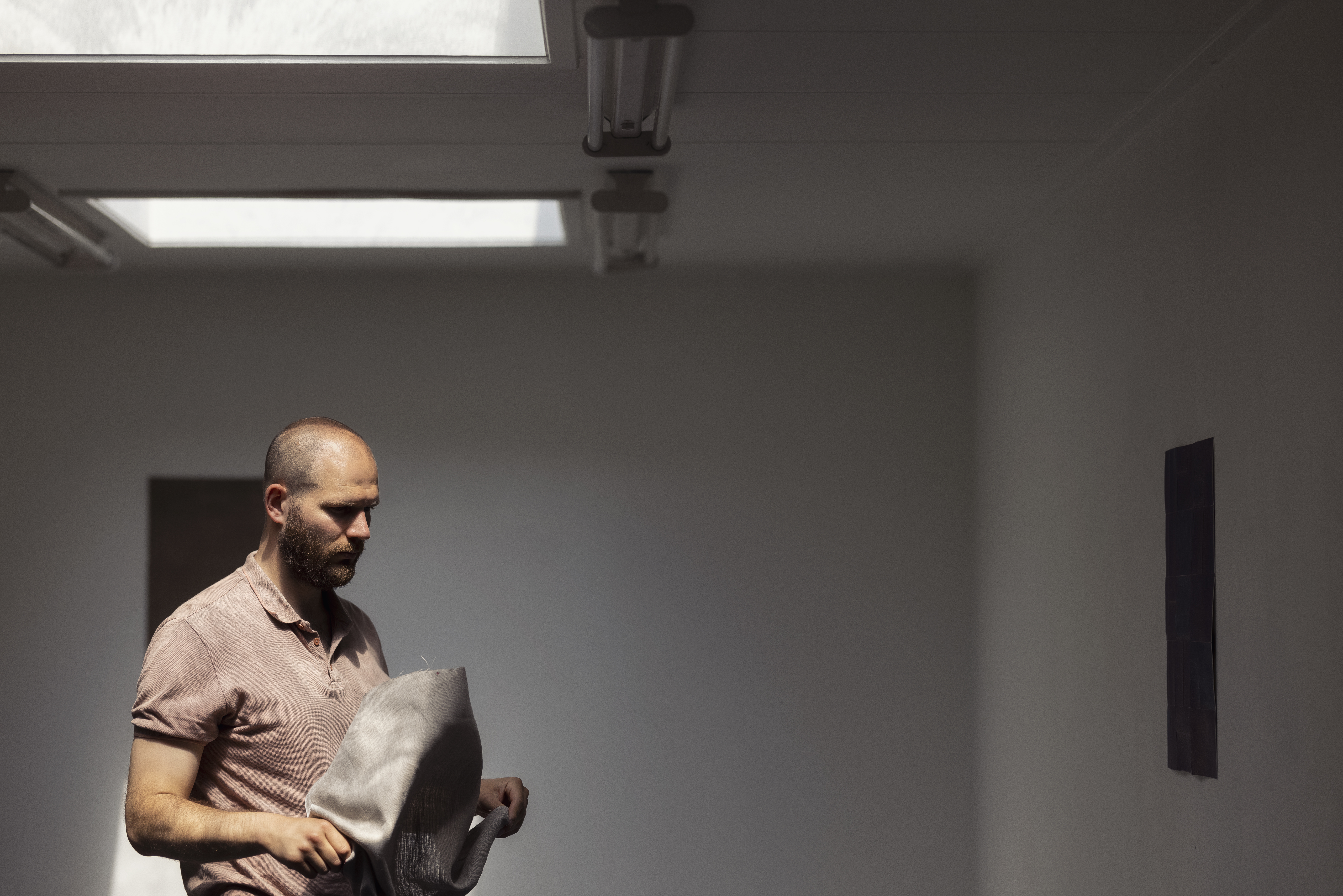 Maarten Dekker staat in het atelier in het licht vanuit de lichtkoepel en houdt een doek in de handen